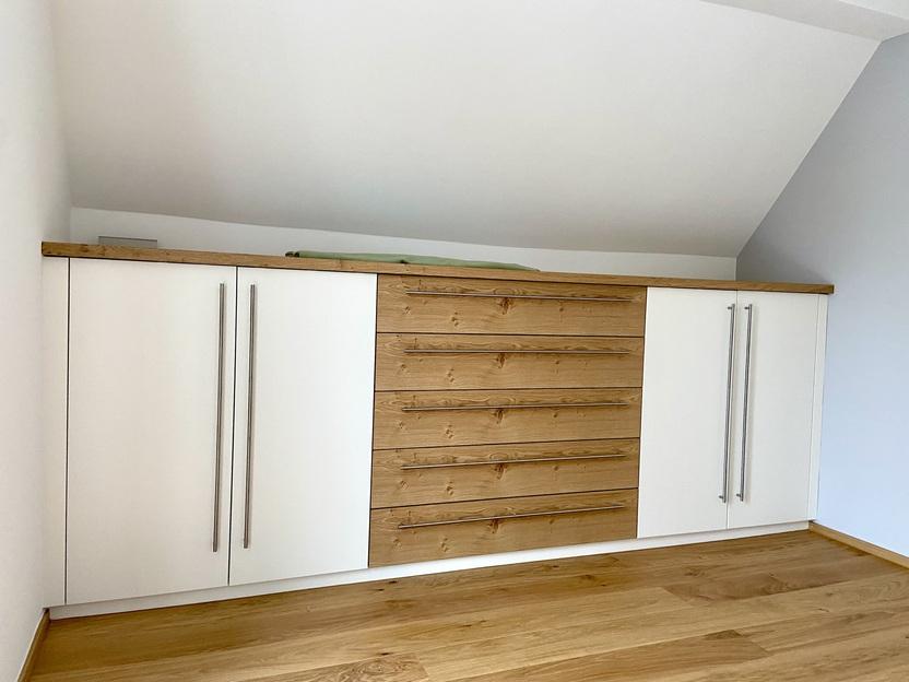 Kommode nach Maß unter Dachschräge Maßgefertigte Kommode aus Holz und Weiß unter einer Dachschräge, mit langen Griffen und Holzdetails in der Mitte. Ideal für Stauraum in Wohnräumen mit begrenzter Höhe.