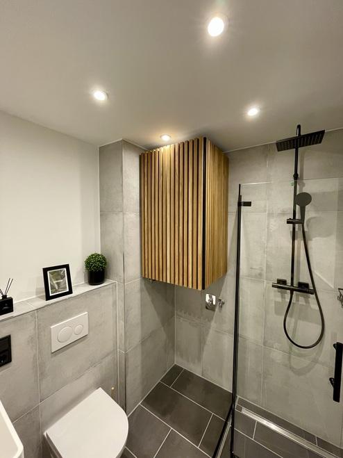 Maßgefertigter Badschrank aus Holz mit vertikalen Lamellen in einem modernen Badezimmer mit grauen Fliesen, Regendusche und eleganter Beleuchtung.