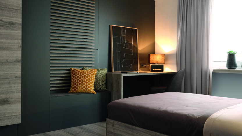 Moderne Schlafzimmeransicht mit einem Bett, Schreibtisch, Sessel, Regalen und Pflanzen vor einem Fenster.